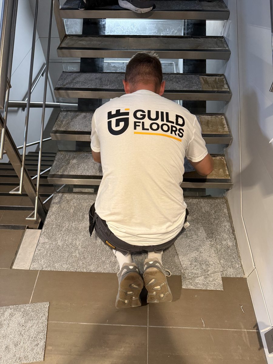 A Guild Floors fitter in a white branded t-shirt laying carpet tiles on a staircase